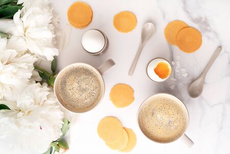 Soft-boiled egg with liquide orange yolk in ceramic egg cups, two cups of coffee with ceramic spoons on background of beautiful white peony. Breakfast concept. Top view.の写真素材