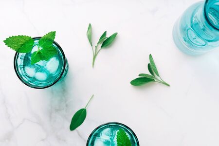 Top view of two beautiful vintage turquoise glasses and a bottle with cold drink and ice cubes, decorated with fresh green mint leaves on white marble table. Copy space.の写真素材