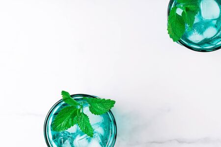 Two beautiful vintage turquoise glasses with cold drink and ice cubes, decorated with fresh green mint leaves on white marble table. Top view.の写真素材
