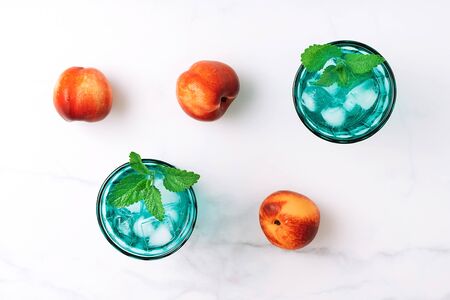 Two beautiful vintage turquoise glasses with cold drink and ice cubes, decorated with fresh green mint leaves and three nectarins on white marble table. Top view.の写真素材