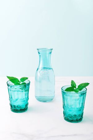 Two beautiful vintage turquoise glasses and a bottle with cold drink and ice cubes, decorated with fresh green mint leaves on white marble table. Copy space.の写真素材