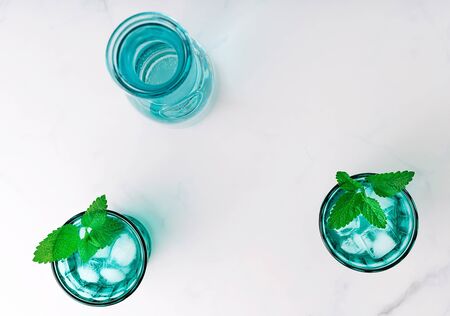 Top view of two beautiful vintage turquoise glasses and a bottle with cold drink and ice cubes, decorated with fresh green mint leaves on white marble table. Copy space.の写真素材
