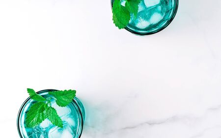 Two beautiful vintage turquoise glasses with cold drink and ice cubes, decorated with fresh green mint leaves on white marble table. Top view.の写真素材