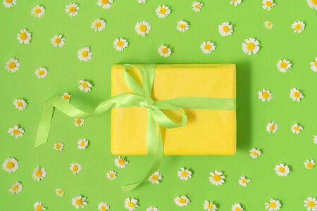 Yellow gift box with green silk ribbon on pastel green background with fresh camomile blossoms. Holiday gift concept. Flat lay. Copy space.の写真素材