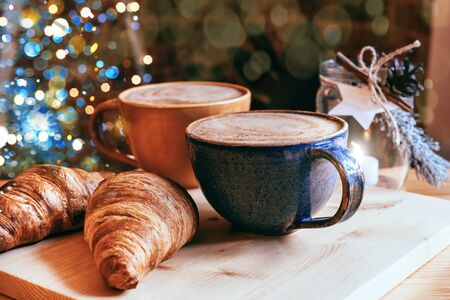 Perfect breakfast of croissants and cappuccino in a cafe. Blue and yellow ceramic cups of cappuccino and croissants on wooden table with Christmas lights and bokeh background. Close-up.の写真素材