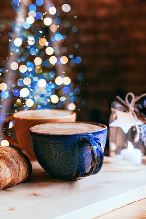 Perfect breakfast of croissants and cappuccino in a cafe. Blue and yellow ceramic cups of cappuccino and croissants on wooden table with Christmas lights and bokeh background. Close-up.の写真素材