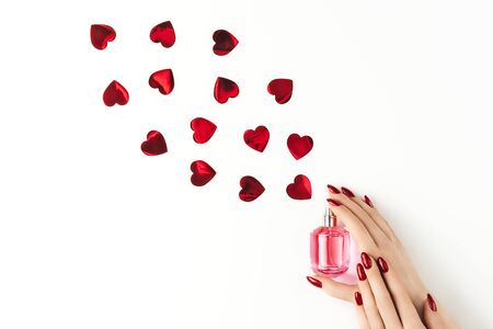 Beautiful female hands with trendy red manicure holding stylish bottle of perfume with spray of vibrant red heart-shaped confetti on white background. Creative trendy flat lay with space for text. Mockup for your design. Love and celebration concept.の写真素材
