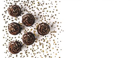 Three delicious chocolate cupcakes with chocolate icing and sprinkled gold sparkles on white background with golden star shaped confetti. Holiday concept. Web banner. Flat lay style. Place for text.の写真素材