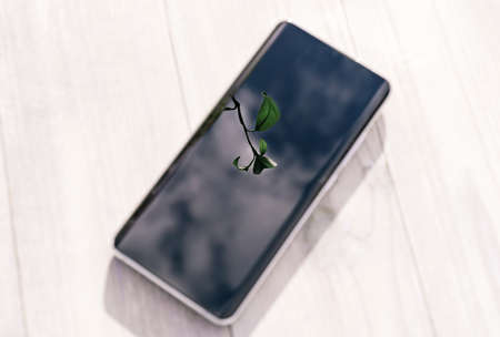Green leaf and sky reflection in smartphone lying on white wooden table. Green energy and modern...