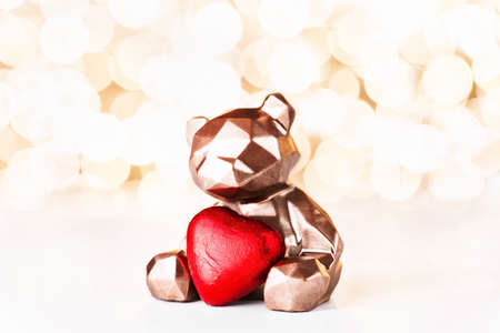 Gift card with chocolate bear holding red heart on golden background of christmas lights bokeh. Mockup for decorative design. Christmas and New Year concept. Copy space.の写真素材