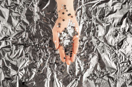 Female hand with trendy silver nail design on foil background holding silver star confetti. Festive backdrop for holidays. Holiday silver manicure. Flat lay style.の写真素材