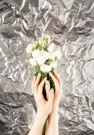 Female hands with trendy silver nail design on silver foil background holding white flowers. Holiday silver manicure. Spring concept. Copy space.の写真素材