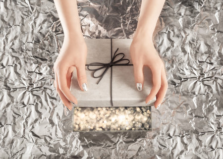 Female hands with beautiful silver nail design opening present box with elegant bow on silver foil background. Christmas lights shining inside the box. Festive backdrop for holiday design. Flat lay style.の写真素材