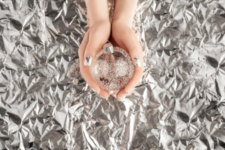 Female hands with trendy silver nail design on foil background holding silver Christmas ball. Festive backdrop for holidays. Holiday silver manicure.の写真素材
