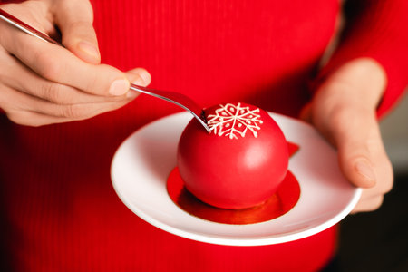 Beautiful young female hands holding red ball mousse dessert decorated with white snowflake on white plate. Christmas celebration concept. Winter season backdrop for your design.の写真素材