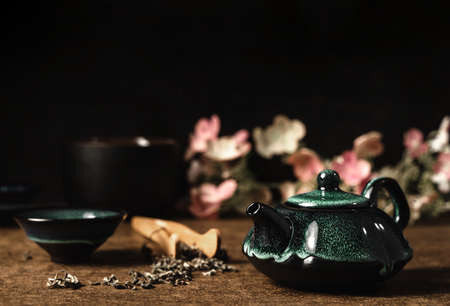 Dark turquoise clay teapot and small cup on wooden background with scattered tea and pink and white...
