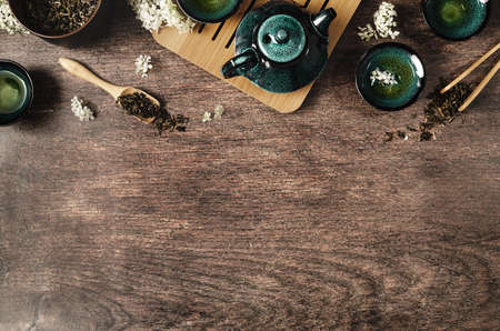 Dark turquoise glazed clay teapot and small cups on dark wooden background decorated with white flowers. Creative minimalist tea ceremony concept. Copy space, top view.の写真素材