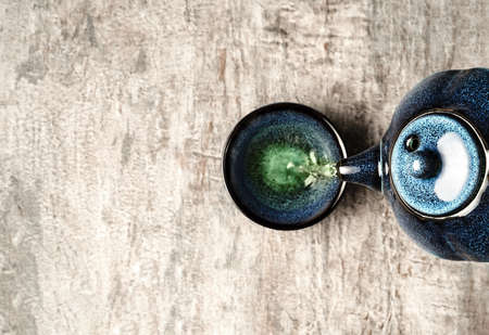 Green tea pouring from a dark turquoise clay teapot in small cup on grey wooden background. Creative minimalist tea ceremony concept. Copy space, top view.の写真素材