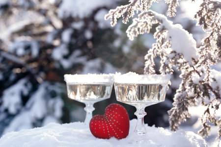 Two crystal glasses of sparkling wine or champagne and red heart in snow on winter natural background with beautiful sunlight bokeh. Love and romance concept.の写真素材