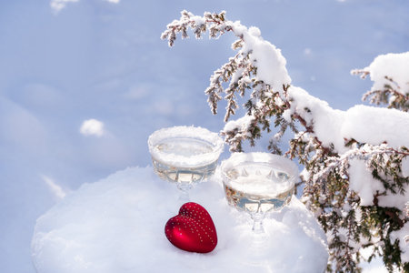 Two crystal glasses of sparkling wine or champagne and red heart in snow on winter natural background. Love and romance concept.の写真素材