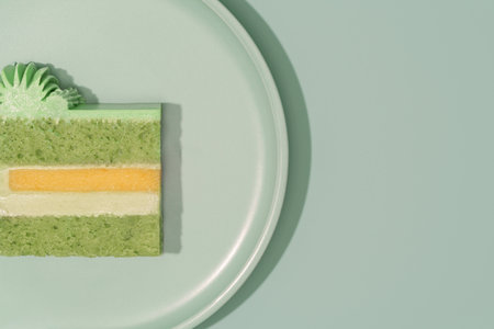 Colorful bento cake slice on a pale green plate, revealing layers of green pistachio, cream, mango,...