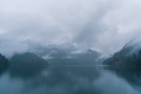 Serene lake with tranquil waters reflects misty mountains and cloudy sky, creating a peaceful atmosphere. The surrounding trees add depth to the landscape, enhancing the natural beauty of the sceneの写真素材