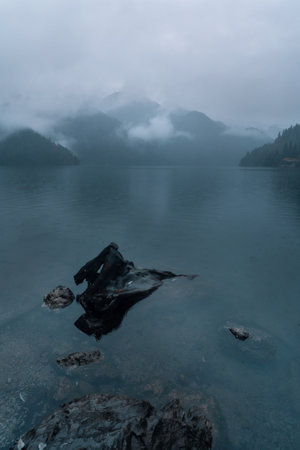 Serene lake landscape features misty mountains in the background and a rocky shoreline, creating a tranquil atmosphere with calm waters reflecting the cloudy sky and surrounding natureの写真素材
