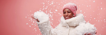African American woman wearing a cozy winter coat and pink hat is joyfully tossing snowflakes in the air, creating a playful winter atmosphere with ample copy space for design elementsの素材