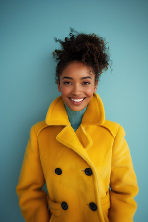 African American woman with curly hair is smiling brightly while wearing a vibrant yellow coat, set against a striking blue background, creating a cheerful and lively atmosphereの素材