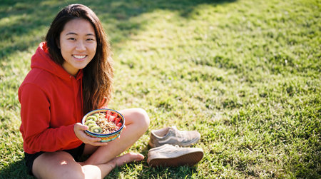 Asian woman dressed in a red hoodie is happily sitting on lush green grass, holding a vibrant bowl filled with assorted fresh fruits, radiating joy and a healthy lifestyleの素材