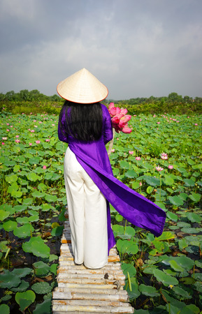 Vietnamese woman wearing traditional dress (ao dai) with conical hat and lotus flowers, standing on wooden bridge.の写真素材