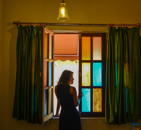 A young woman standing at colorful glass window with curtain in dark room.の写真素材