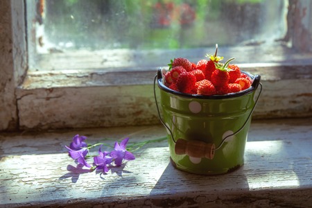 A full small bucket of freshly picked wild strawberries and flowers of bells stands on the windowsill of an abandoned house. Rustic style. The concept of summer, rest, pleasure, healthy eating.の写真素材