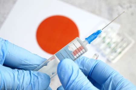 syringe with a covid-19 vaccine is held by hand in a glove on background of the Japanese flag,...