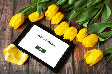 tablet pc and big yellow tulips flowers bouquet with bow on wooden background, florist online with big flowers bunch, workplace florist, closeupの写真素材