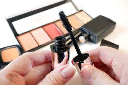 Mascara in hand on set of cosmetic makeup products background. Lipstick, eye shadows, brush, powder,...