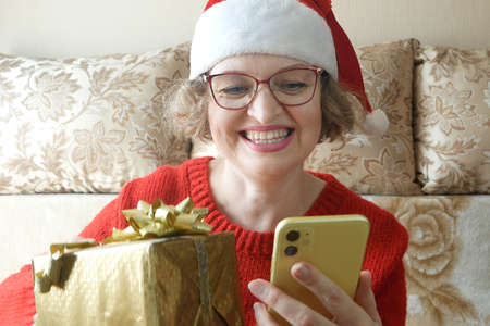 positive woman in santa claus hat use smartphone and box gift send christmas congratulation for friends, relatives, parents in house with x-mas comfort atmosphere indoorsの写真素材