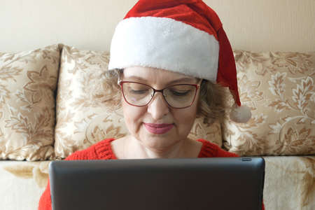 positive woman in santa claus hat use laptop send christmas congratulation for friends, relatives, parents in house with x-mas comfort atmosphere indoorsの写真素材