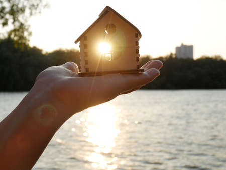 hand hold wooden house model agaist blue sunny sky, forest and river, conceptの写真素材