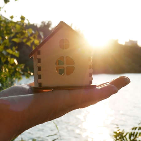 hand hold wooden house model agaist blue sunny sky, forest and river, conceptの写真素材