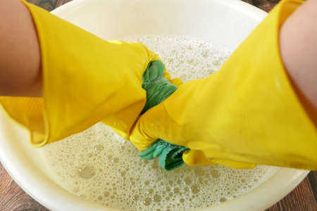 woman hand in yellow gloves rolling out linen in basin, female hands washing color clothes in basin,...