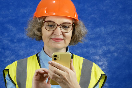 Feamle Engineer in the construction helmet and yellow vest with mobile phone and documents on blue background, closeupの写真素材