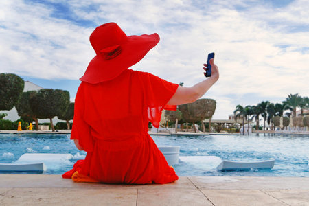 Summer vacation woman in red hat and cape for the beach with mobile phone near swimmimg pool amd palms, woman blogger, outdoorsの写真素材