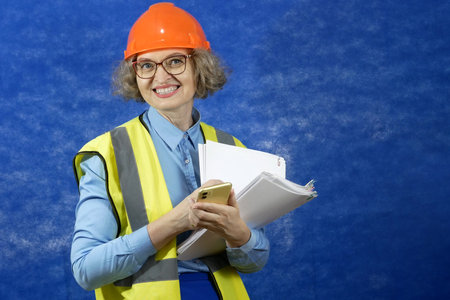 Feamle Engineer in the construction helmet and yellow vest with mobile phone and documents on blue...