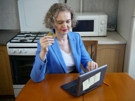 middle aged woman in jacket working at kitchen with laptop and bank card, home office concept, indoorの写真素材