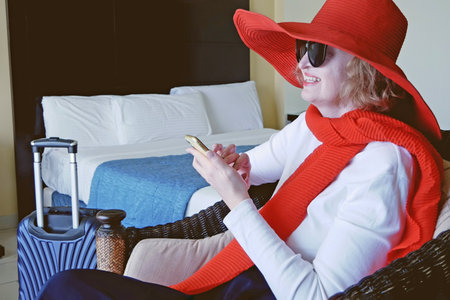 Woman wearing the big red hat with suitcase checking out of hotel with her baggage sitting on bed waiting for taxi ordered on mobile phone app, indoorsの写真素材