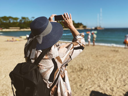 Senior adventureisageless woman with backpack taking a selfie on a mobile phone on the sea beach. Summer active tourism for retirees. Internet holiday conceptの写真素材