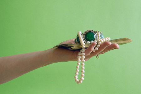 Hand holding vintage silver bracelet with green stones, pearls necklace and golden jewelry green background, closeupの写真素材