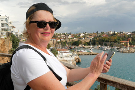 Senior tourist woman consulting gps on smart phone in a touristic place with sea beach Turkey Antalya old town the background, outdoorsの写真素材