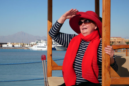 Happy senior woman in red hat and sunglasses waiting your a cruise ship in marina, adventure is less, travel for pensioners, copyspace, outdoorsの写真素材